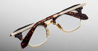 A pair of Jacques Marie Mage Sartre glasses with gold metal rims and tortoiseshell-patterned frames, engraved temples, by Jacques Marie Mage, photographed on a light gray background.