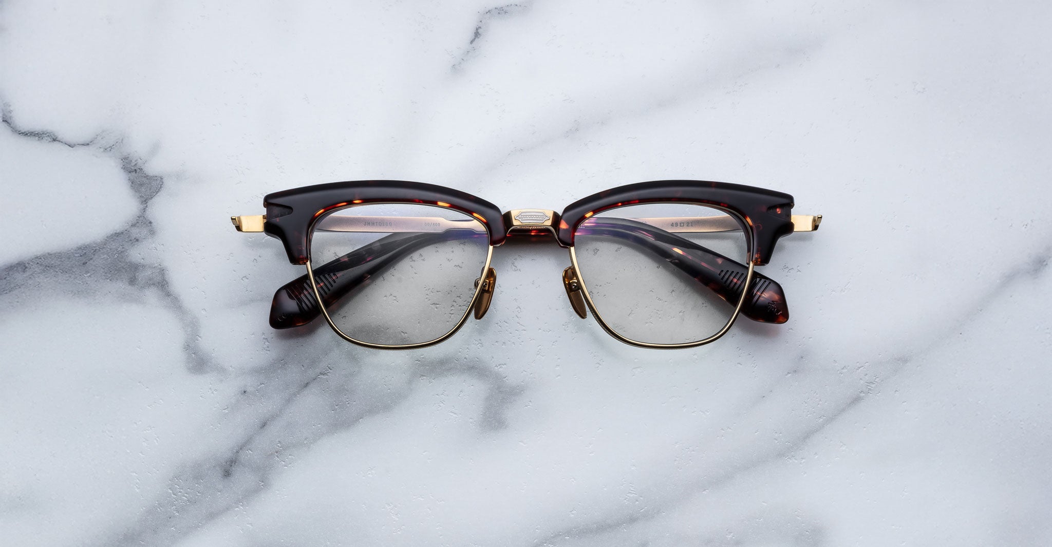 A pair of Jacques Marie Mage Sartre glasses with dark, thick frames and gold accents rests on a white marble surface.
