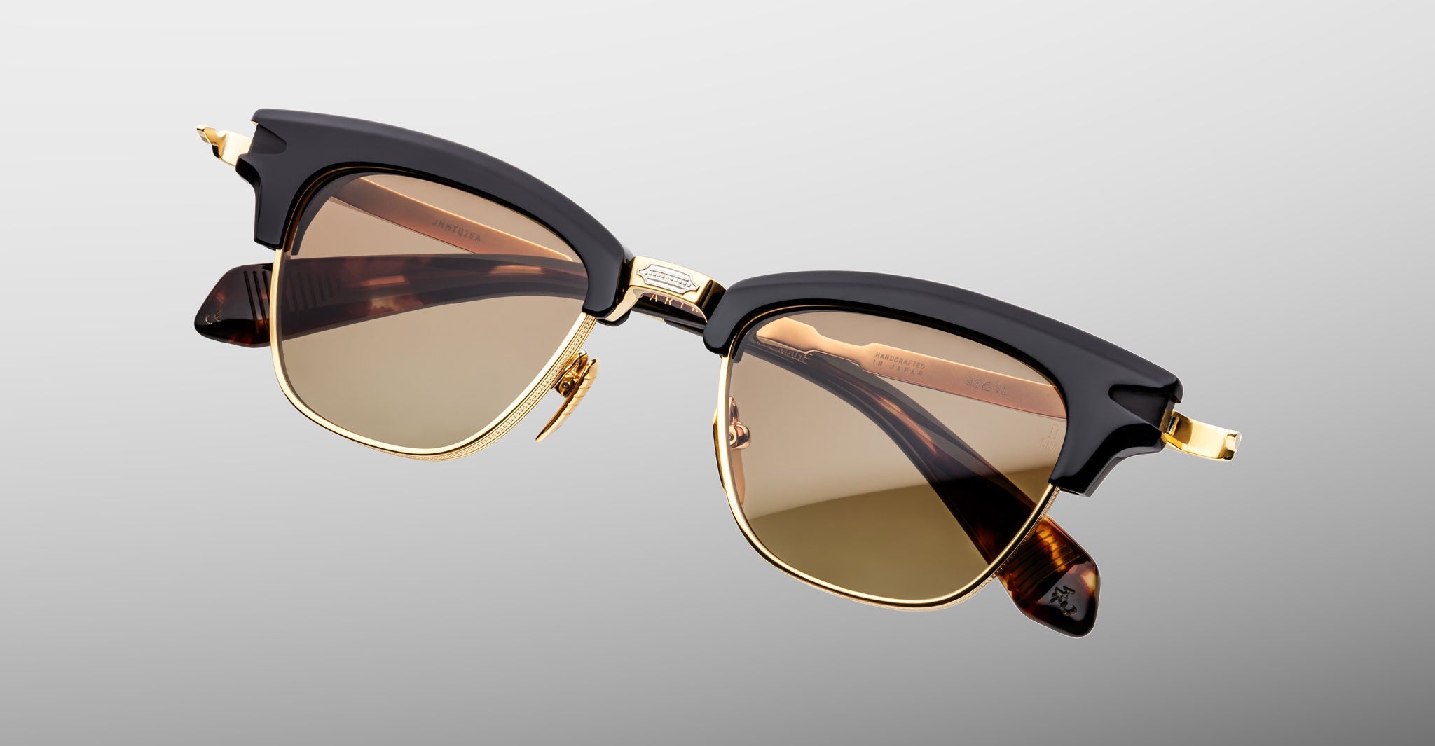 The Jacques Marie Mage Sartre by Jacques Marie Mage are browline sunglasses with gold metal frames, black upper rims, and brown tinted lenses, inspired by classic clubmaster design and shown on a light gray background.