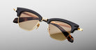 The Jacques Marie Mage Sartre by Jacques Marie Mage are browline sunglasses with gold metal frames, black upper rims, and brown tinted lenses, inspired by classic clubmaster design and shown on a light gray background.