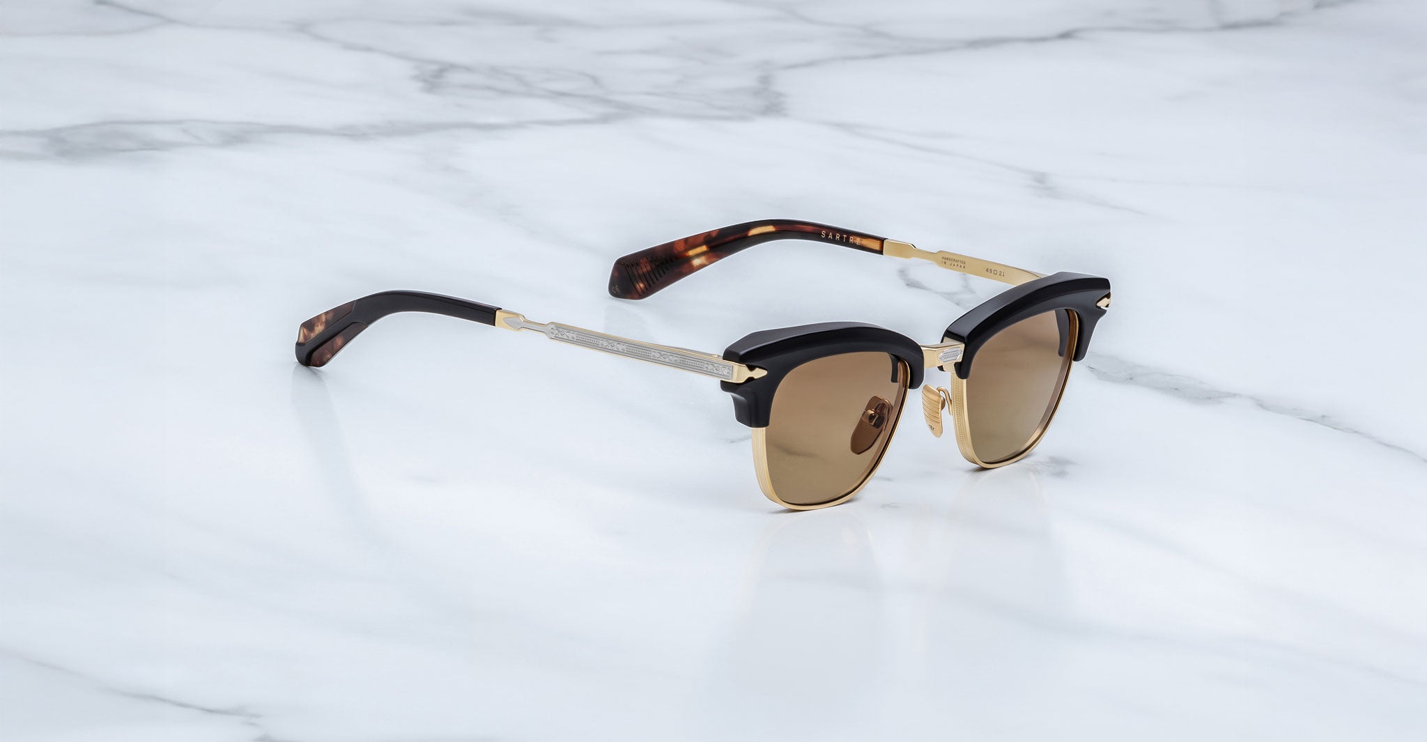 A pair of Jacques Marie Mage Sartre sunglasses with gold and black clubmaster frames and brown lenses rests on a white marble surface.
