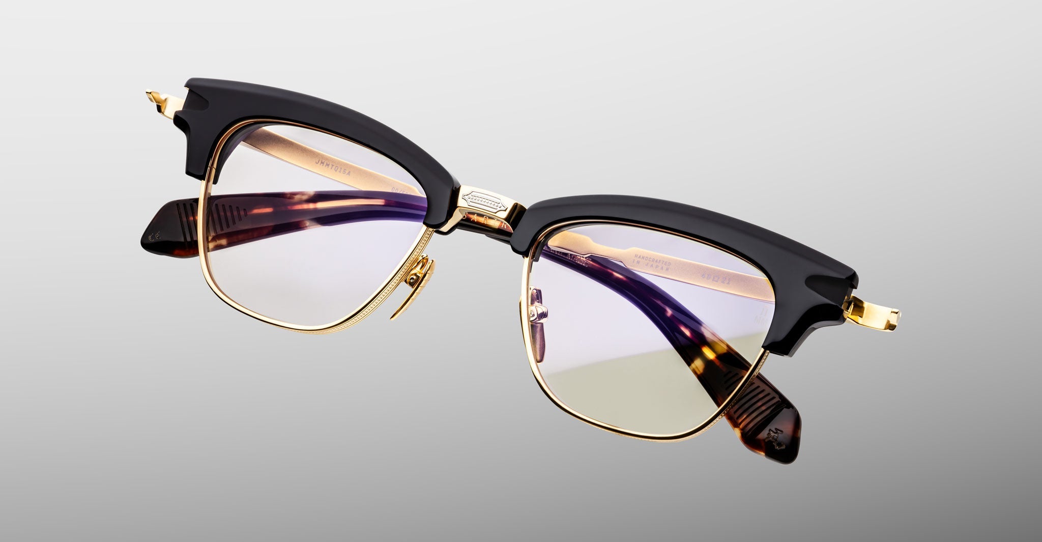 The Jacques Marie Mage Sartre by Jacques Marie Mage features 50s-inspired clubmaster style, black upper rims, a gold titanium frame, and clear lenses, displayed on a light gray background.
