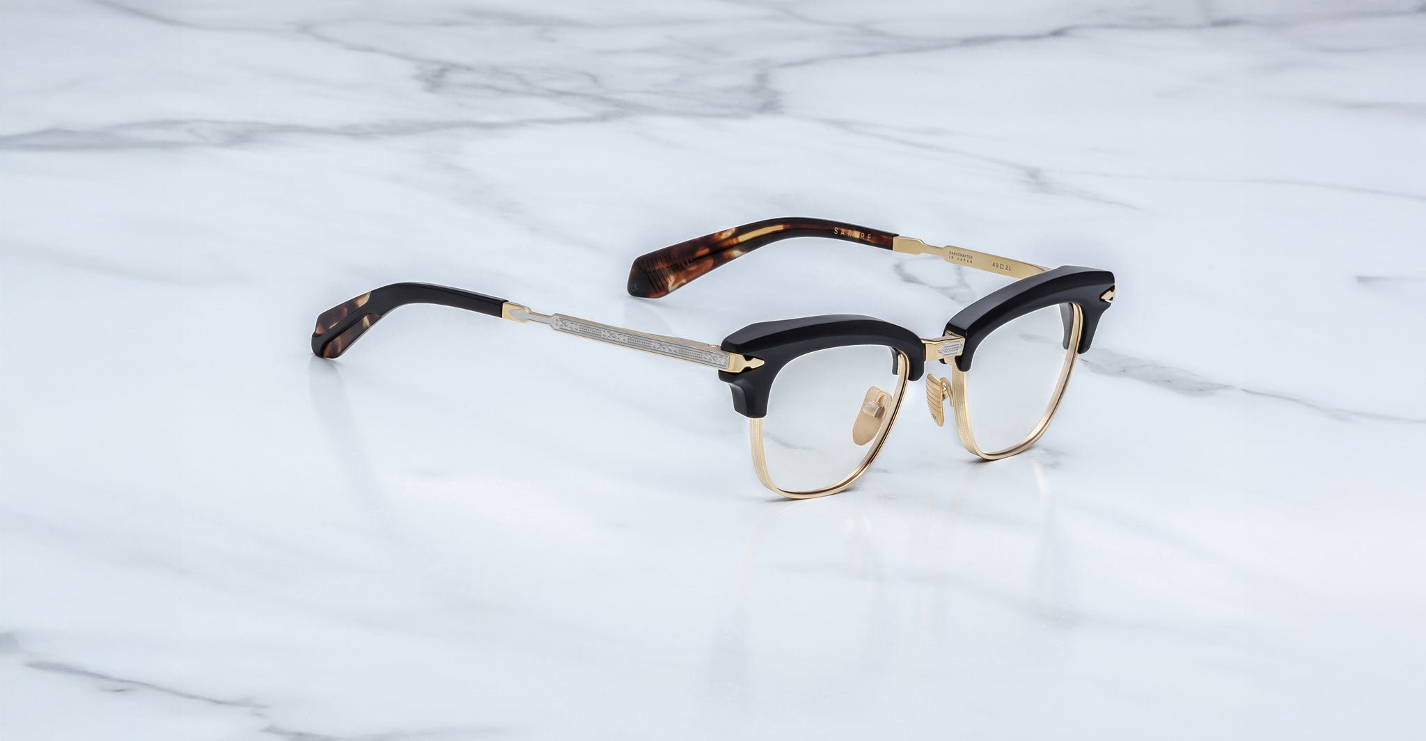 A pair of Jacques Marie Mage Sartre glasses, featuring a black and gold clubmaster frame with tortoiseshell-patterned temple tips, displayed on a white marble surface.