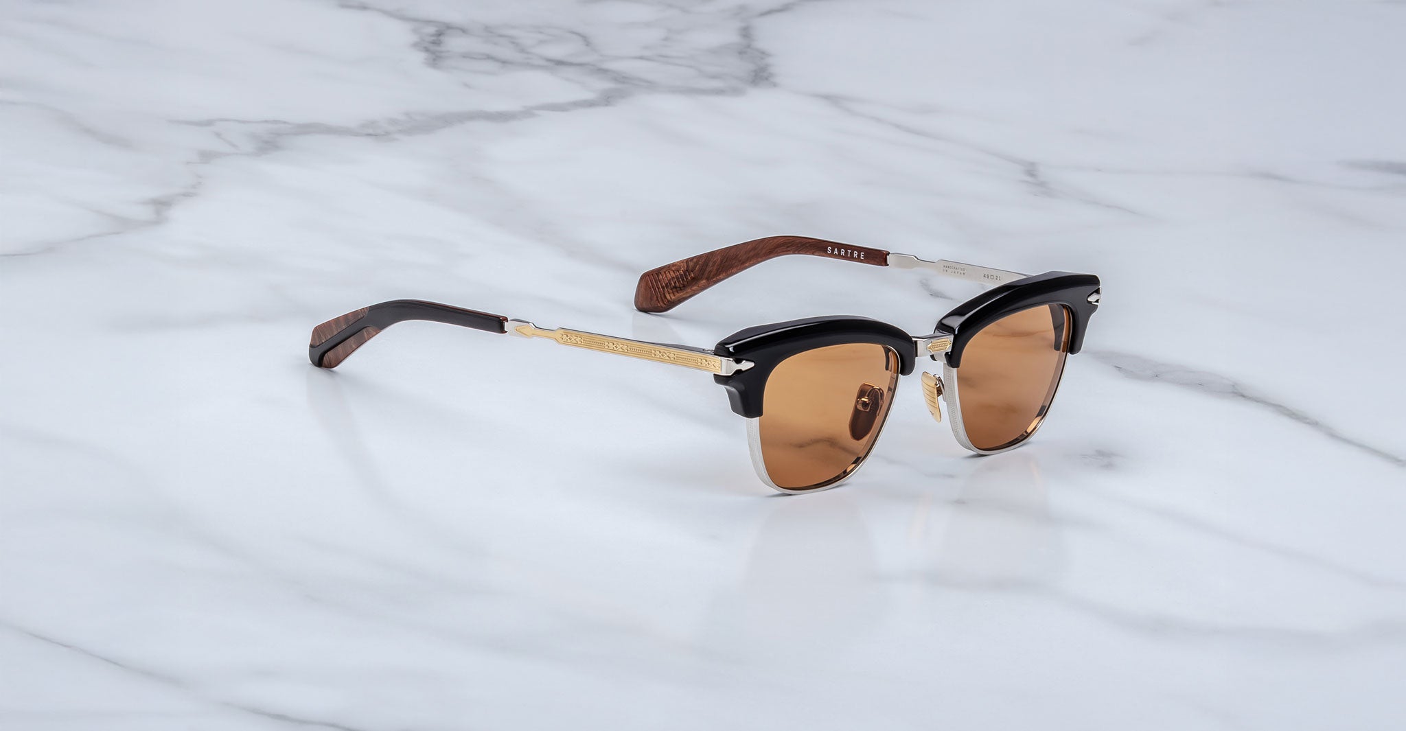 The Jacques Marie Mage Sartre sunglasses by Jacques Marie Mage feature clubmaster frames with brown-tinted lenses, black and gold accents, and wooden arms, elegantly displayed on a white marble surface.