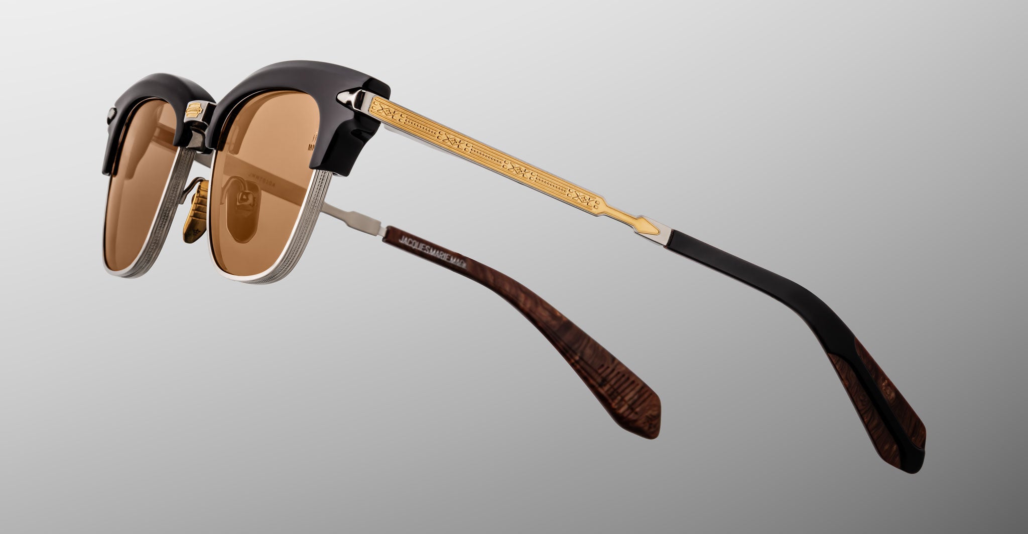 The Jacques Marie Mage Sartre sunglasses by Jacques Marie Mage feature brown lenses, black clubmaster-style upper frames, gold textured temples, and brown-patterned earpieces, shown on a gray background.