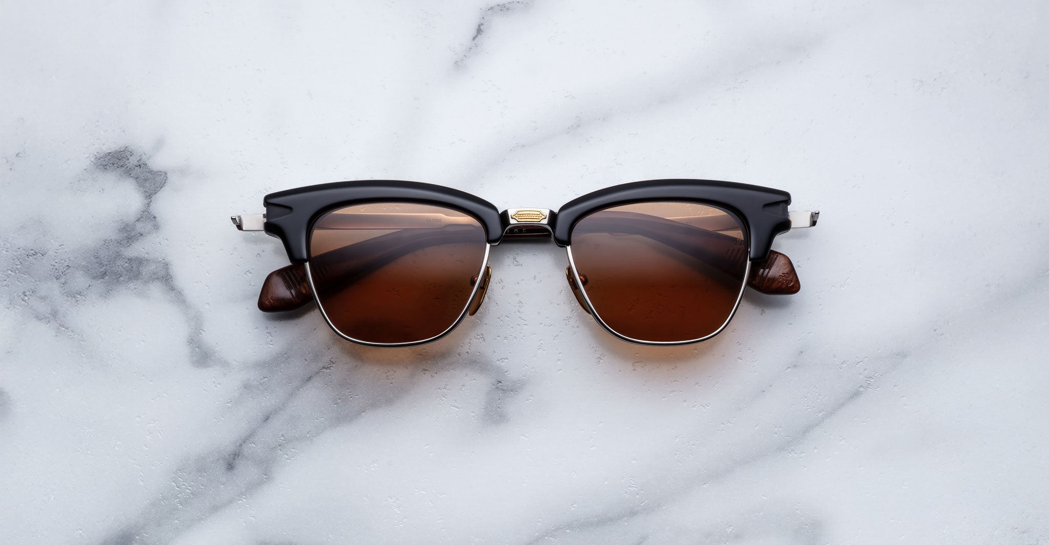 The Jacques Marie Mage Sartre glasses by Jacques Marie Mage, in black and brown with gold accents, rest elegantly on a white marble surface.
