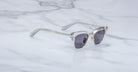 The Jacques Marie Mage Sartre sunglasses by Jacques Marie Mage, featuring clear frames and dark lenses inspired by the iconic clubmaster design, are displayed on a white marble surface.