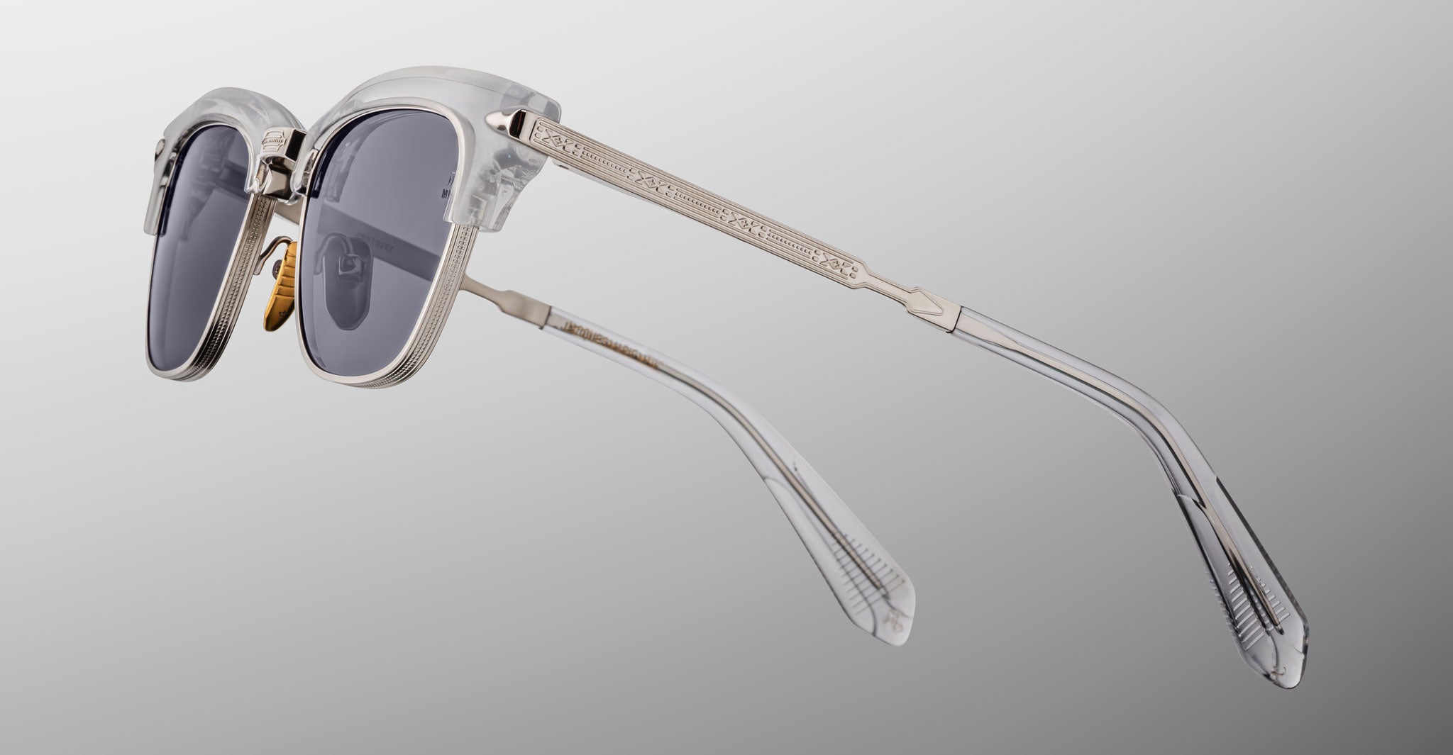 Jacques Marie Mage Sartre sunglasses feature clear frames, dark lenses, and gold-toned metal arms, capturing classic style by Jacques Marie Mage, displayed on a light gray gradient background.
