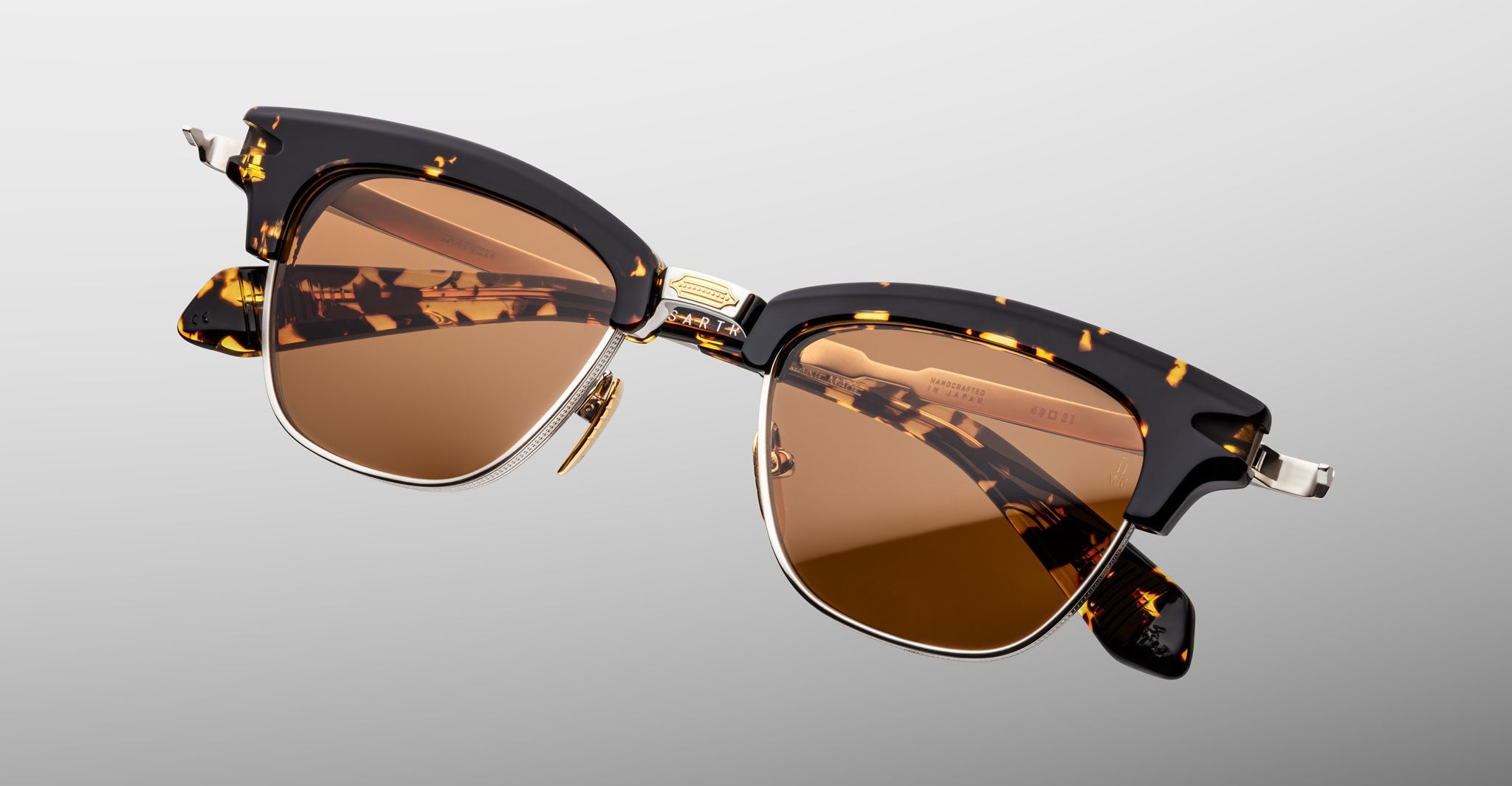 The Jacques Marie Mage Sartre sunglasses from Jacques Marie Mage feature tortoiseshell acetate frames with gold accents and brown lenses, inspired by 50s clubmaster style, shown against a light gray background.