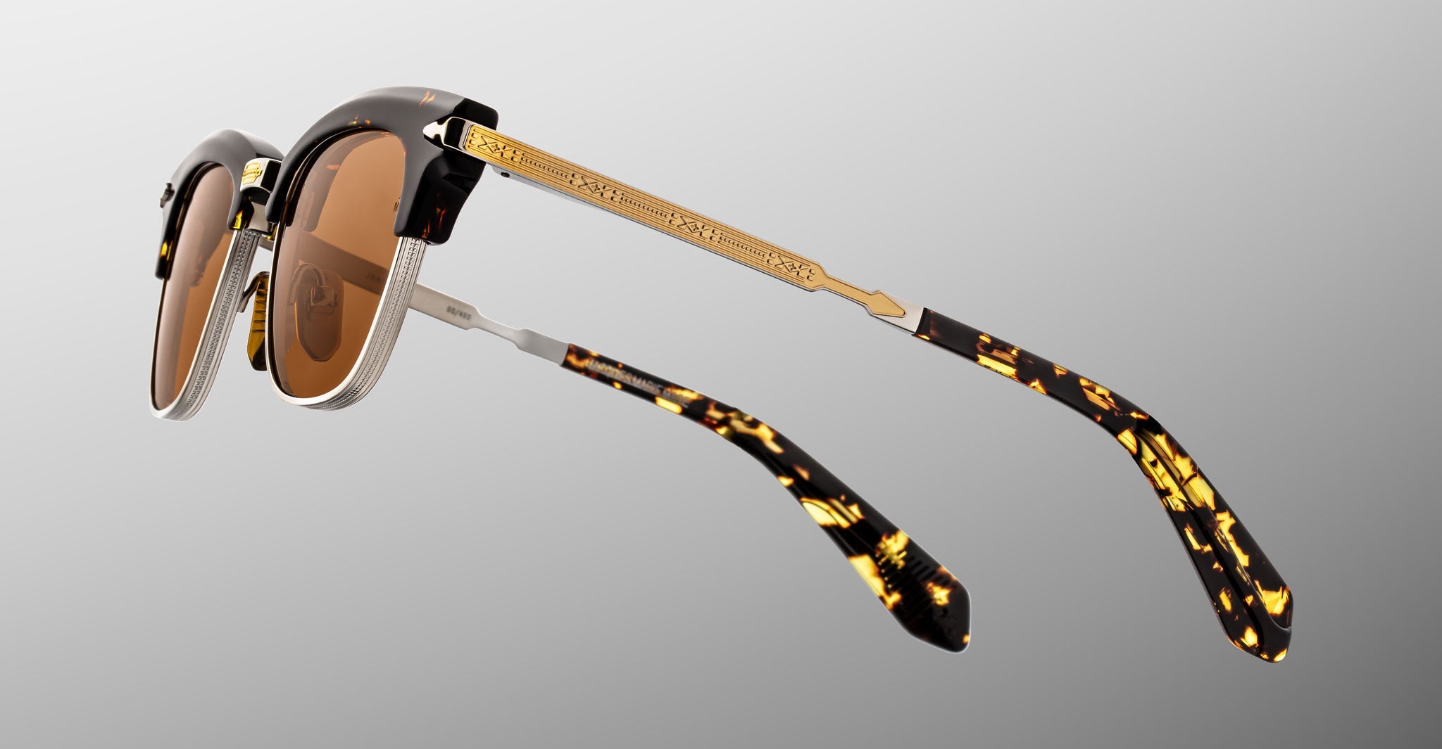 The Jacques Marie Mage Sartre glasses feature brown lenses, gold metal arms with etched detailing, and tortoiseshell-patterned tips in a clubmaster style, shown on a light gray background.