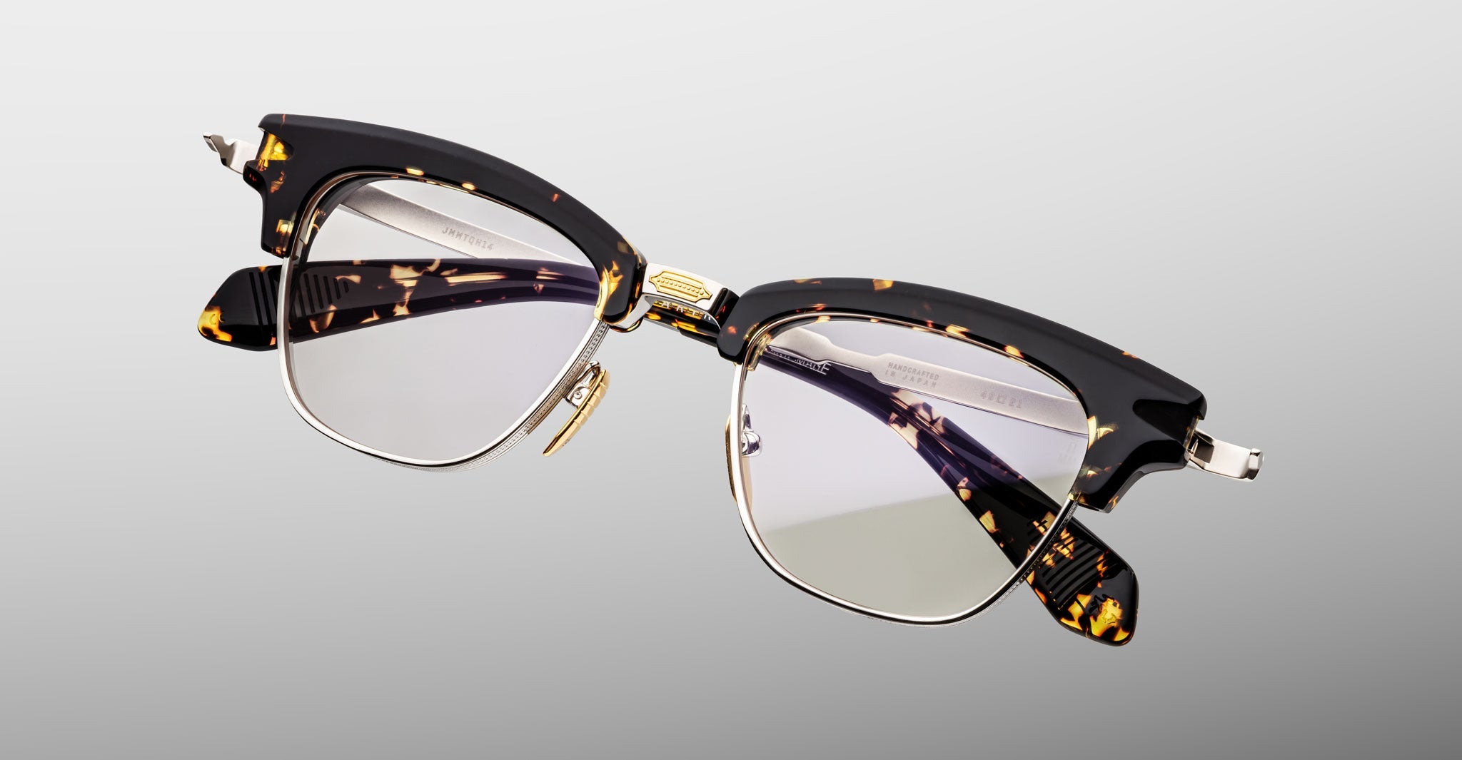 The Jacques Marie Mage Sartre by Jacques Marie Mage are clubmaster eyeglasses in tortoiseshell and black with clear lenses, gold detailing, and metal accents, displayed on a light gray background.