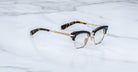 Jacques Marie Mage Sartre optical eyeglasses by Jacques Marie Mage, featuring gold metal rims and black tortoiseshell arms, are displayed on a white marble surface.