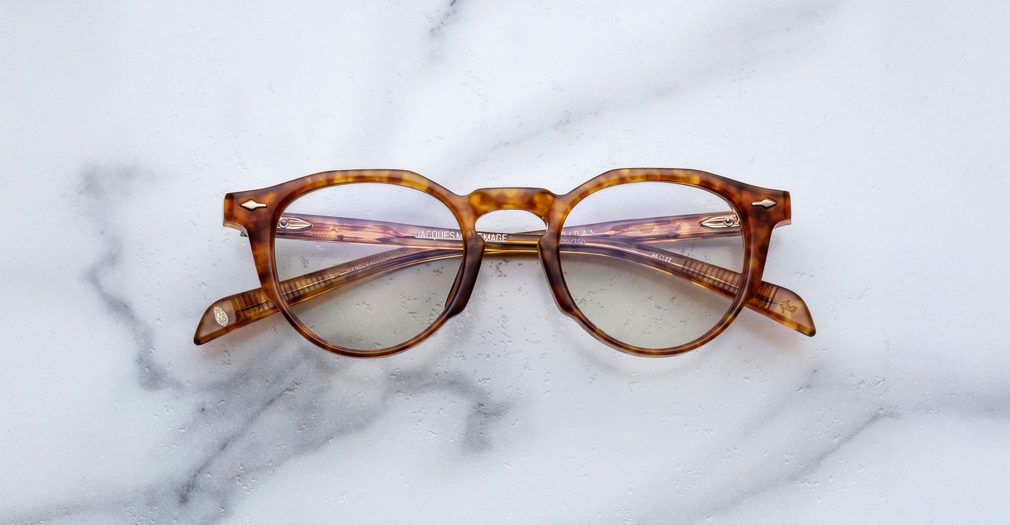 Jacques Marie Mage Sheridan round tortoiseshell eyeglasses with clear lenses displayed on a white marble surface, arms folded outward to reveal frame details.