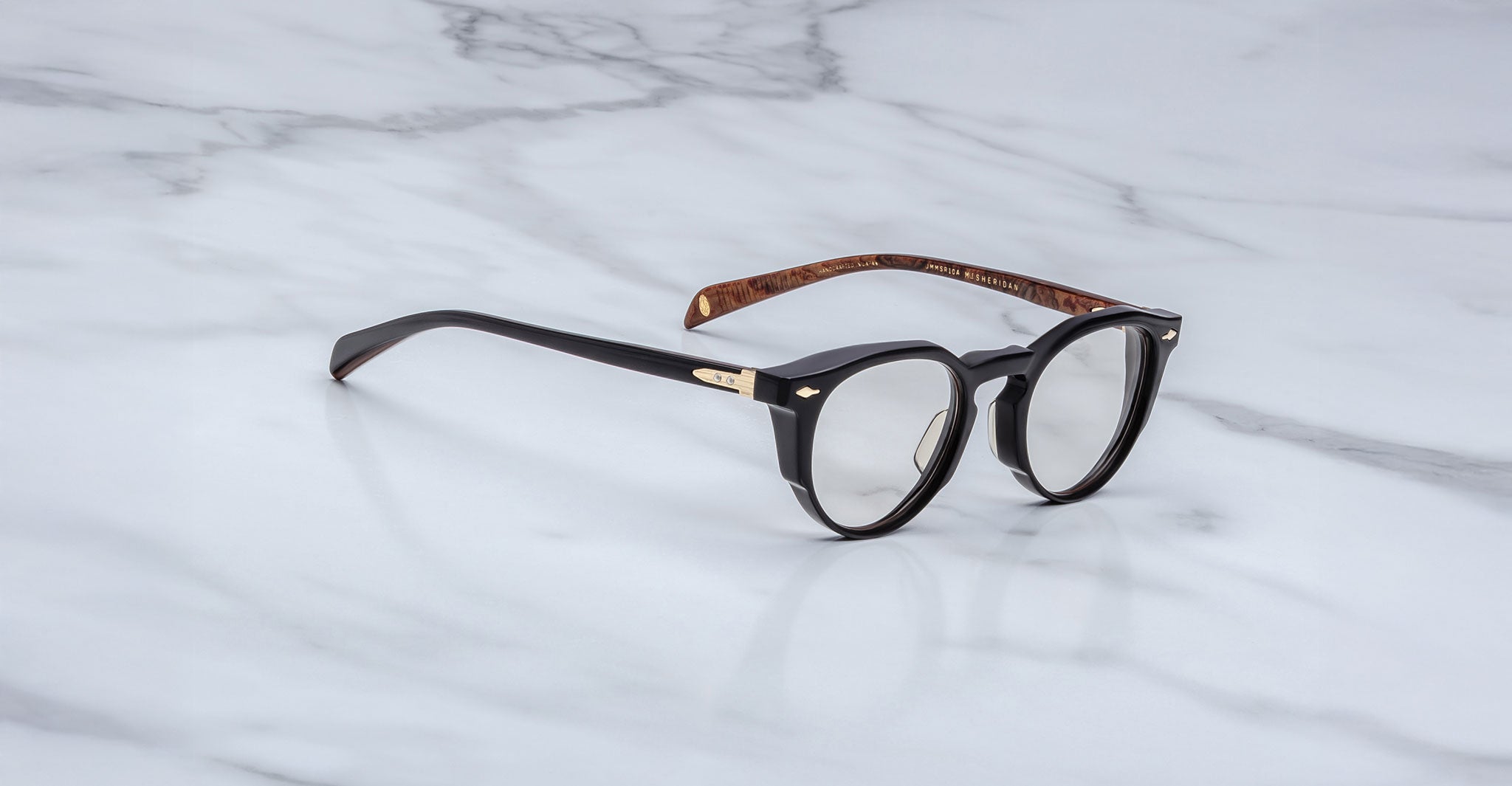 A pair of Jacques Marie Mage Sheridan round black eyeglasses with gold hinge detailing and brown tortoiseshell inner arms rests on a white marble surface.