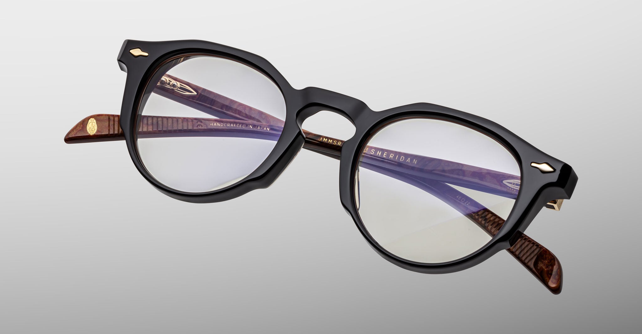 The Jacques Marie Mage Sheridan features round eyeglasses with thick black frames, brown textured temple arms, and clear lenses. Logo details appear on the inner arms, all set against a light gray background.