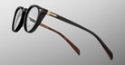 The Jacques Marie Mage Sheridan eyeglasses are shown in close-up, featuring black frames with gold accents and brown patterned inner temples, set against a gray gradient background.