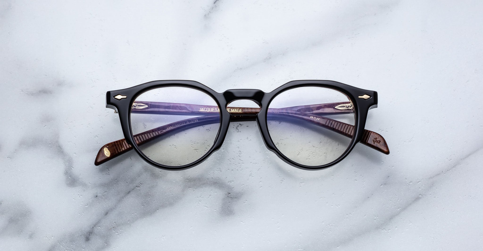 A pair of Jacques Marie Mage Sheridan round black eyeglasses with brown arms is placed on a white marble surface.