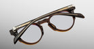 The Jacques Marie Mage Sheridan eyeglasses, round and brown in acetate with metal hinges, are shown folded lens-side down on a light gray background.