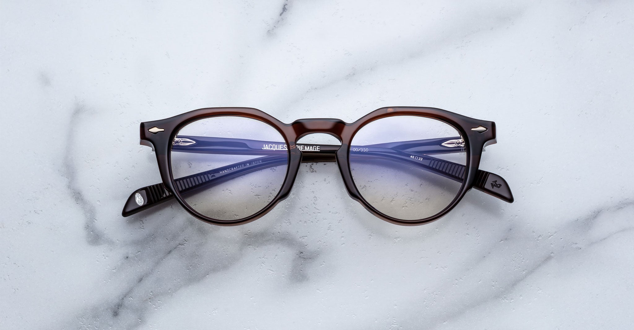 The Jacques Marie Mage Sheridan eyeglasses, featuring round dark brown frames with blue lenses and folded arms, rest on a white marble surface.