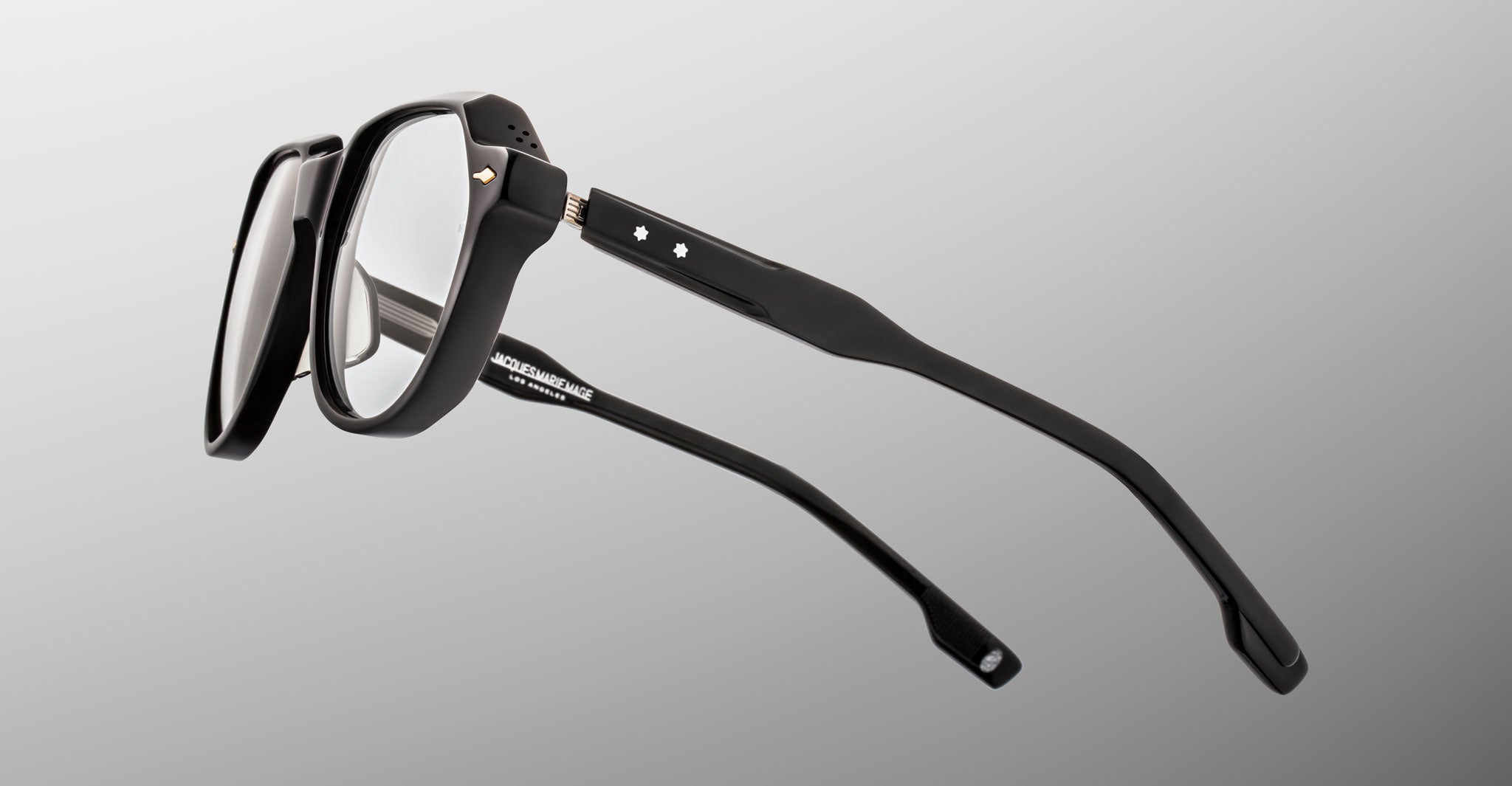 The Jacques Marie Mage Shozo eyeglasses feature black thick-rimmed frames with clear lenses, viewed from an angled side perspective on a light gray background. Two small white star accents adorn the temples.