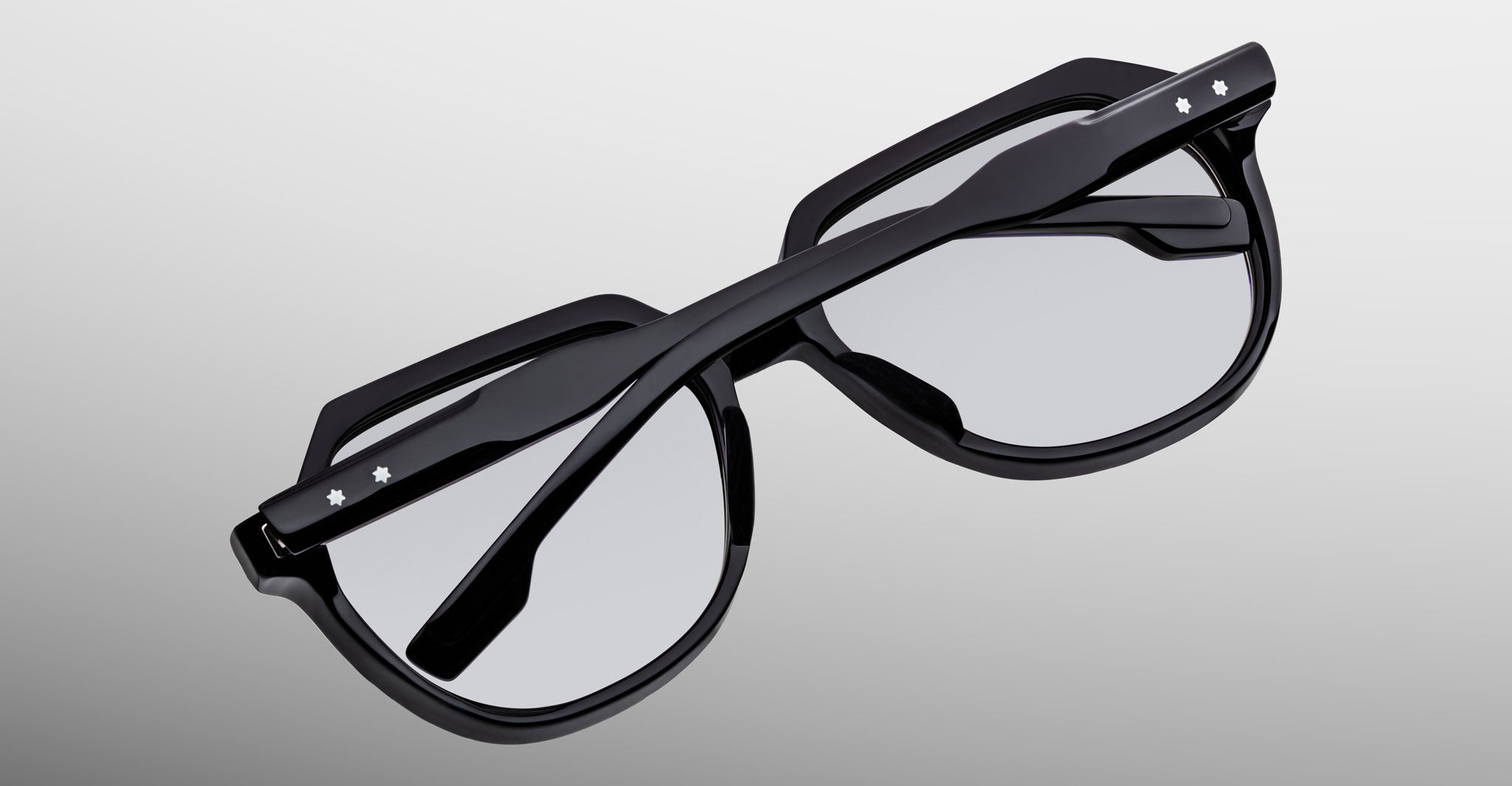 The Jacques Marie Mage Shozo glasses, black with thick rims and folded arms, feature small white star-shaped accents on the arms and are shown from above against a light gray gradient background.