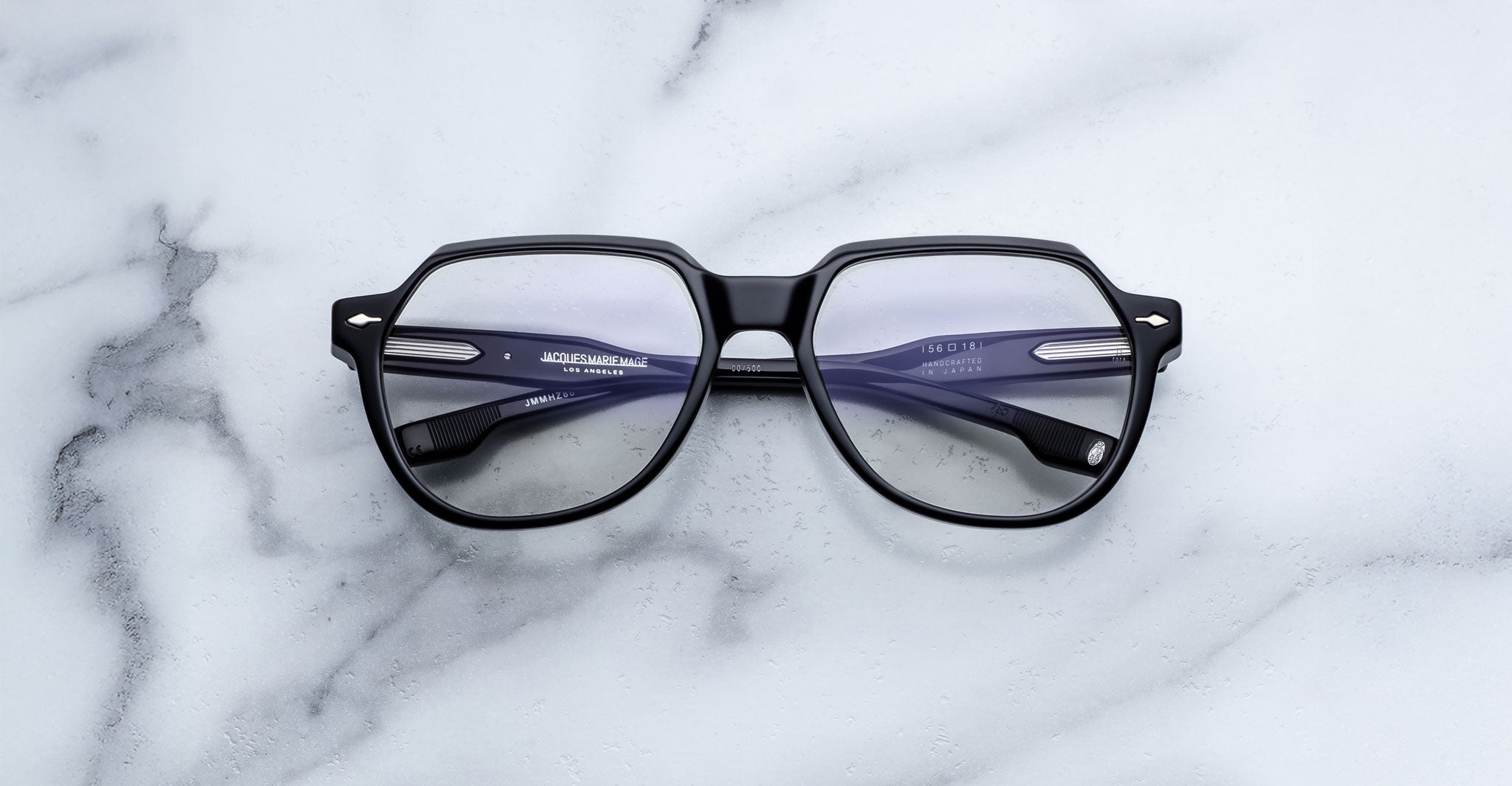 The Jacques Marie Mage Shozo eyeglasses, featuring black frames and clear lenses, rest on a white marble surface. The temples display brand and size details, and the frames have a subtle shine.