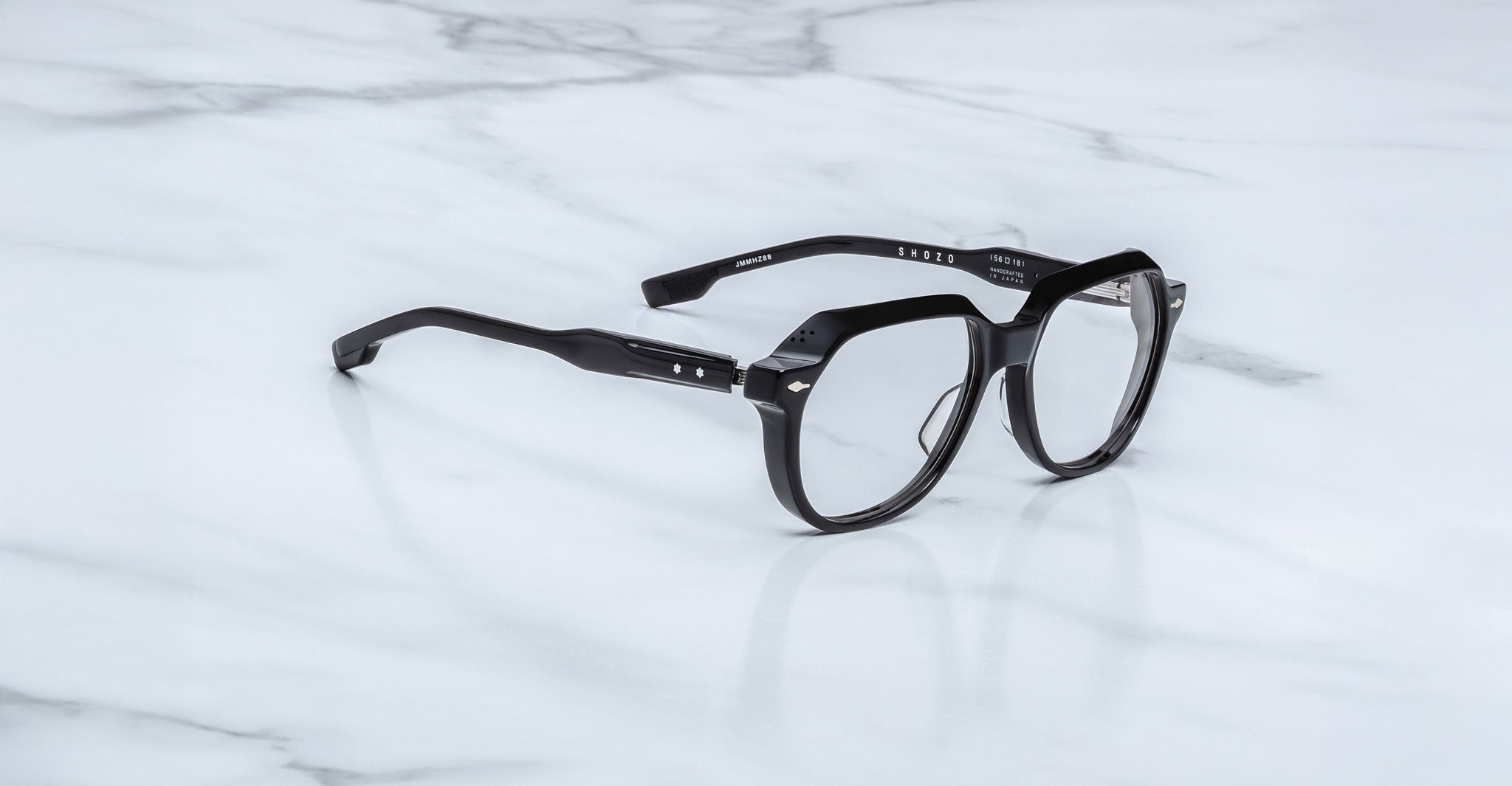 Jacques Marie Mage Shozo eyeglasses with clear lenses and thick black frames rest on a white marble surface. The arms are slightly bent, featuring metallic accents near the hinges.
