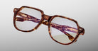 The Jacques Marie Mage Shozo is a pair of stylish, square-shaped eyeglasses with tortoiseshell frames and clear lenses, shown against a plain light gray background. The temples feature distinct detailing and engraved text.