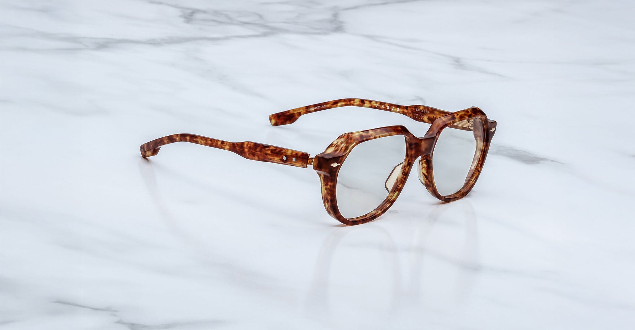 A pair of Jacques Marie Mage Shozo geometric brown tortoiseshell eyeglasses rests on a white marble surface.