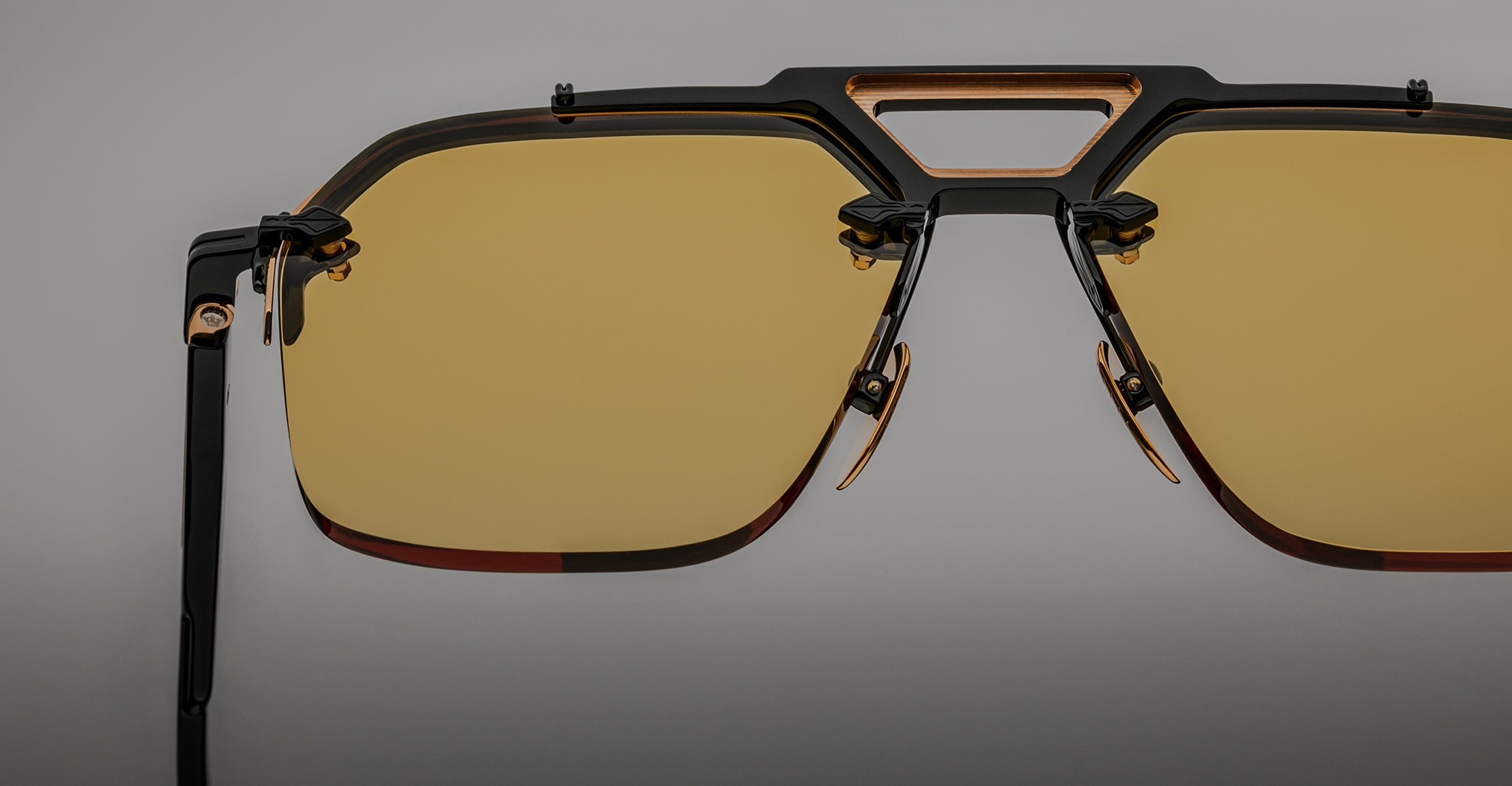 Close-up of Jacques Marie Mage Silverton sunglasses featuring yellow-tinted lenses, a geometric metal frame with black and gold accents, and adjustable nose pads on a neutral gray background.