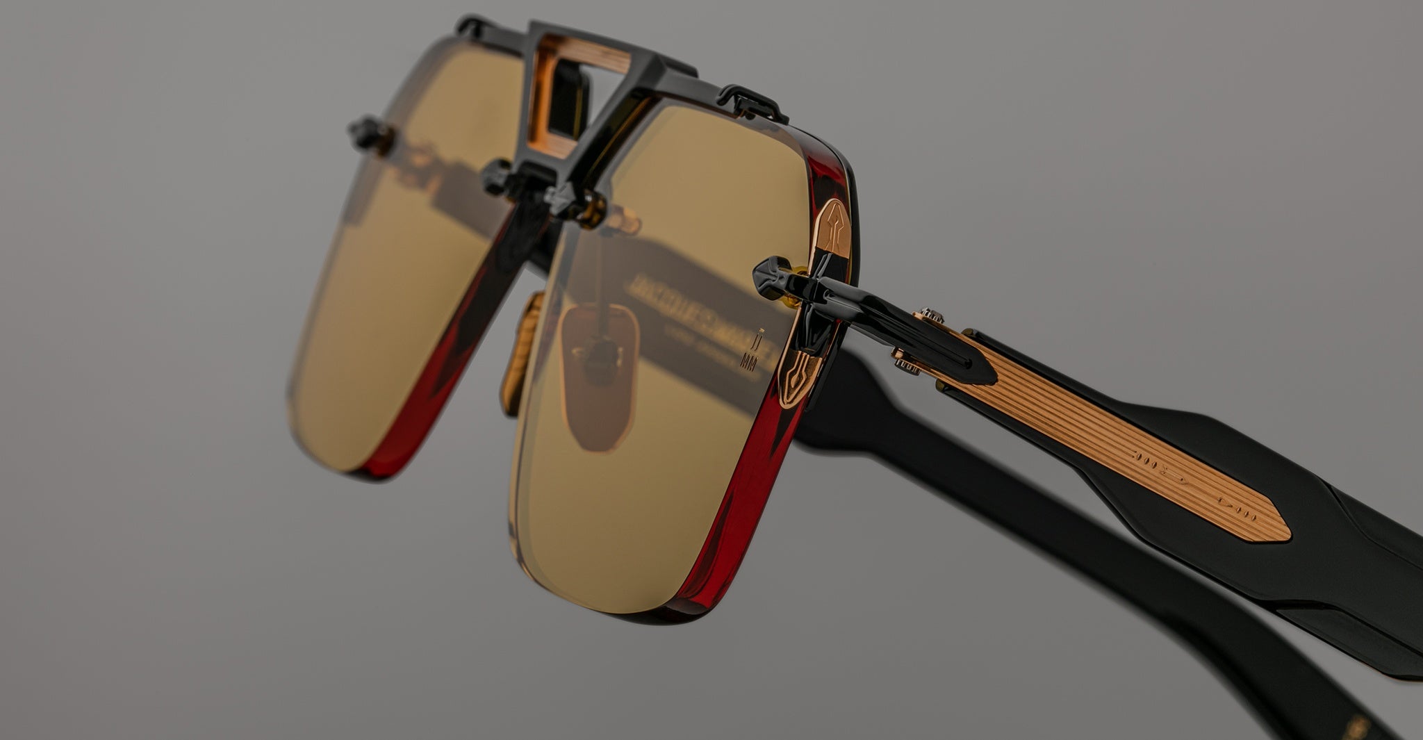 Close-up of Jacques Marie Mage Silverton sunglasses featuring rectangular yellow-tinted lenses, black and red frames, and metallic gold accents on the arms, set against a plain gray background.