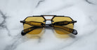 A pair of Jacques Marie Mage Silverton black sunglasses with yellow-tinted lenses rests on a white marble surface. Gold brand markings and text appear on the inner sides of the arms.