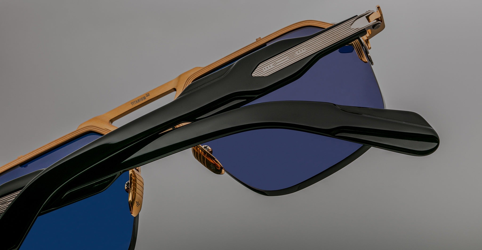Close-up view of Jacques Marie Mage Silverton sunglasses featuring gold frames, blue-tinted lenses, and black arms, set against a plain gray background.