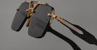 Close-up of Jacques Marie Mage Silverton sunglasses with dark rectangular lenses, gold metal frame, and black arms with metallic detailing, set against a plain gray background.