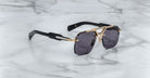 The Jacques Marie Mage Silverton sunglasses feature dark rectangular lenses, gold-tone metal frames, and thick black arms, displayed on a white marble surface with grey veining.