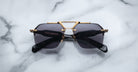 The Jacques Marie Mage Silverton sunglasses, featuring dark lenses and a gold metal geometric frame, are displayed on a white marble surface.