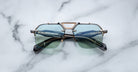 The Jacques Marie Mage Silverton sunglasses feature a geometric gold and black frame with blue-tinted lenses, showcased on a white marble surface.