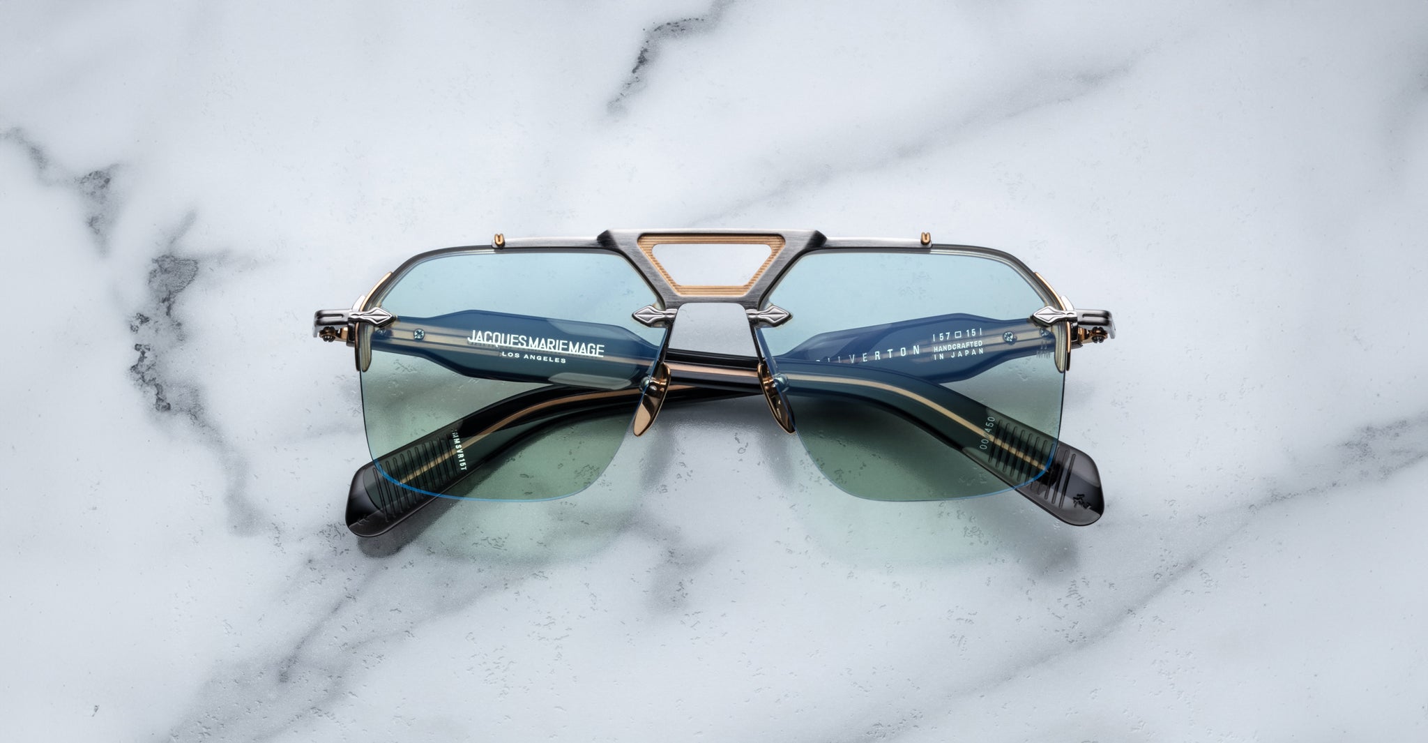 The Jacques Marie Mage Silverton sunglasses feature a geometric gold and black frame with blue-tinted lenses, showcased on a white marble surface.