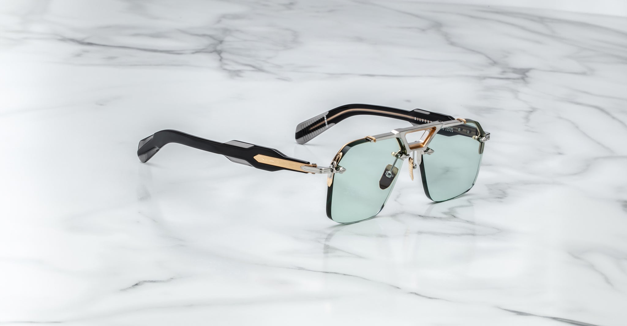 Jacques Marie Mage Silverton sunglasses, featuring green-tinted square lenses, gold and silver metal frame accents, and black arms, rest on a white marble surface.