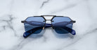 A pair of Jacques Marie Mage Silverton sunglasses with blue lenses and geometric metal accents on the bridge rests upside down on a white marble surface.
