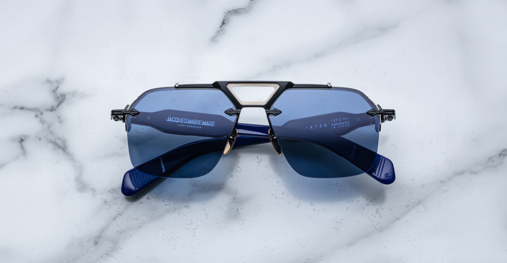 A pair of Jacques Marie Mage Silverton sunglasses with blue lenses and geometric metal accents on the bridge rests upside down on a white marble surface.