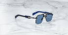 The Jacques Marie Mage Silverton sunglasses, featuring blue lenses, a metallic frame, and blue-black temples, are displayed on a white marble surface.