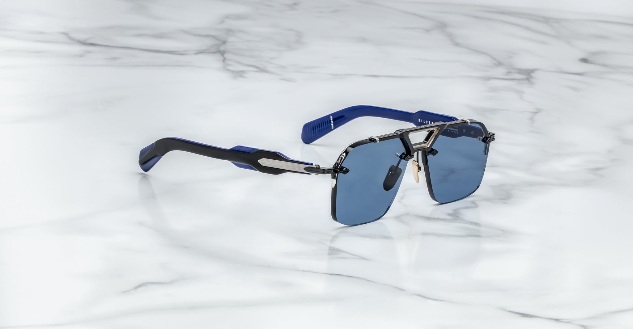 The Jacques Marie Mage Silverton sunglasses, featuring blue lenses, a metallic frame, and blue-black temples, are displayed on a white marble surface.