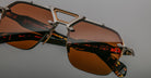 Close-up of Jacques Marie Mage Silverton sunglasses featuring brown tinted lenses, a unique geometric metal frame, and red text branding with specifications on the inner arm. Set against a neutral gray background.