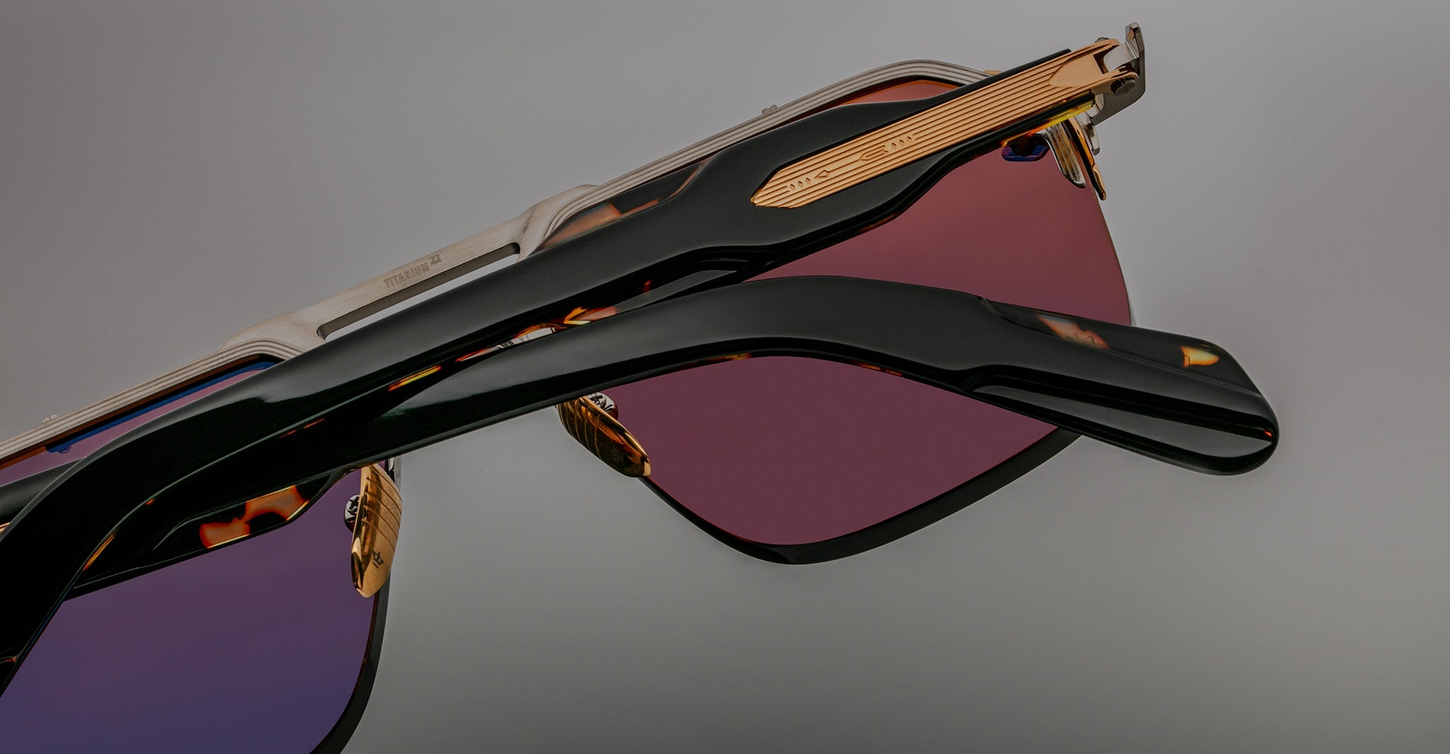 Close-up of two pairs of Jacques Marie Mage Silverton sunglasses with tinted lenses—one purple, one rose—featuring black and metallic gold frames, set against a plain background.