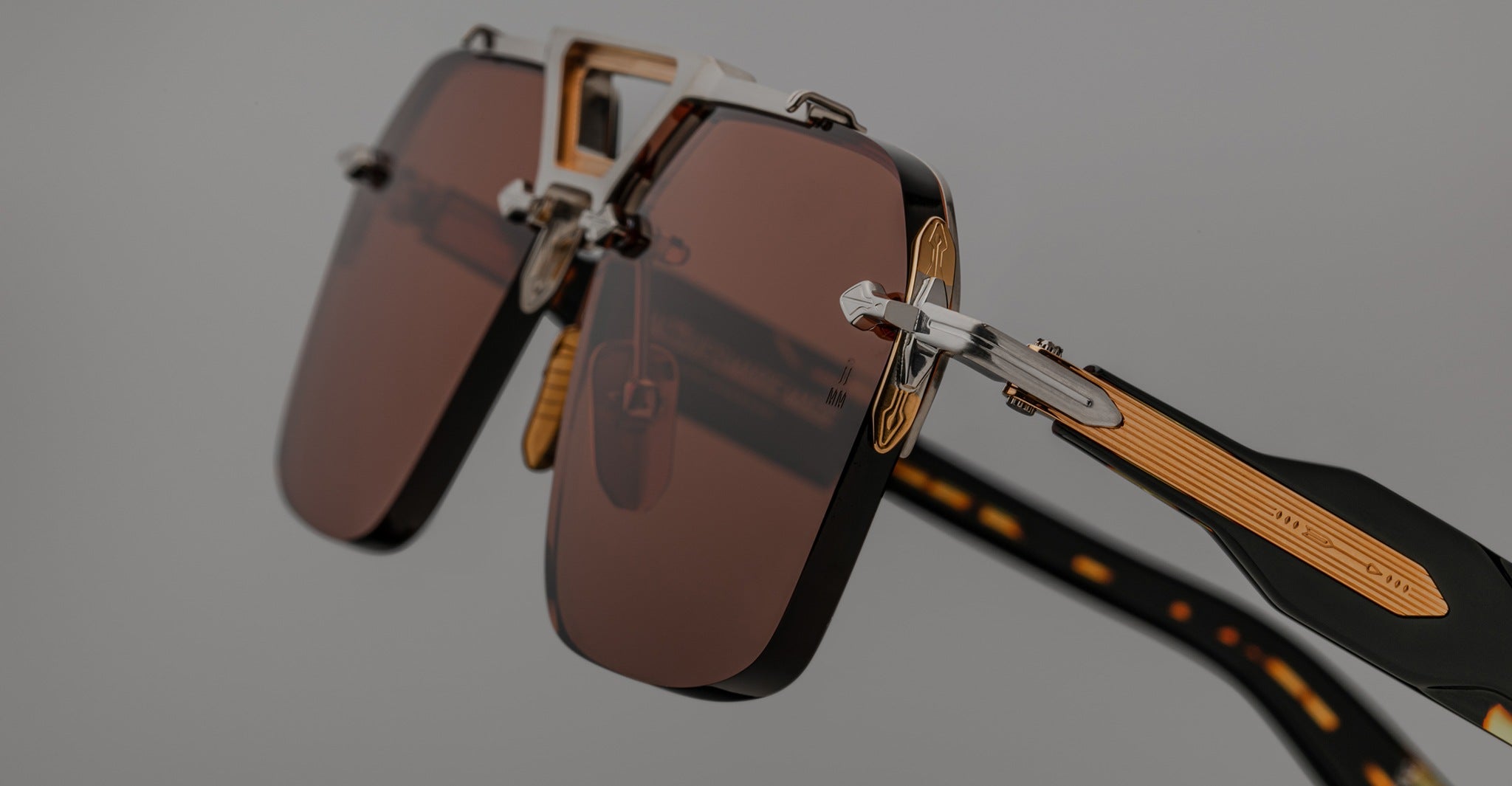 Close-up view of Jacques Marie Mage Silverton sunglasses featuring brown rectangular lenses, gold accents, and patterned black arms, set against a neutral background.