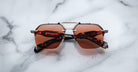 The Jacques Marie Mage Silverton rectangular sunglasses, featuring brown tinted lenses and a silver metal frame, rest on a white marble surface. The brand name and details appear in red text on the arms.