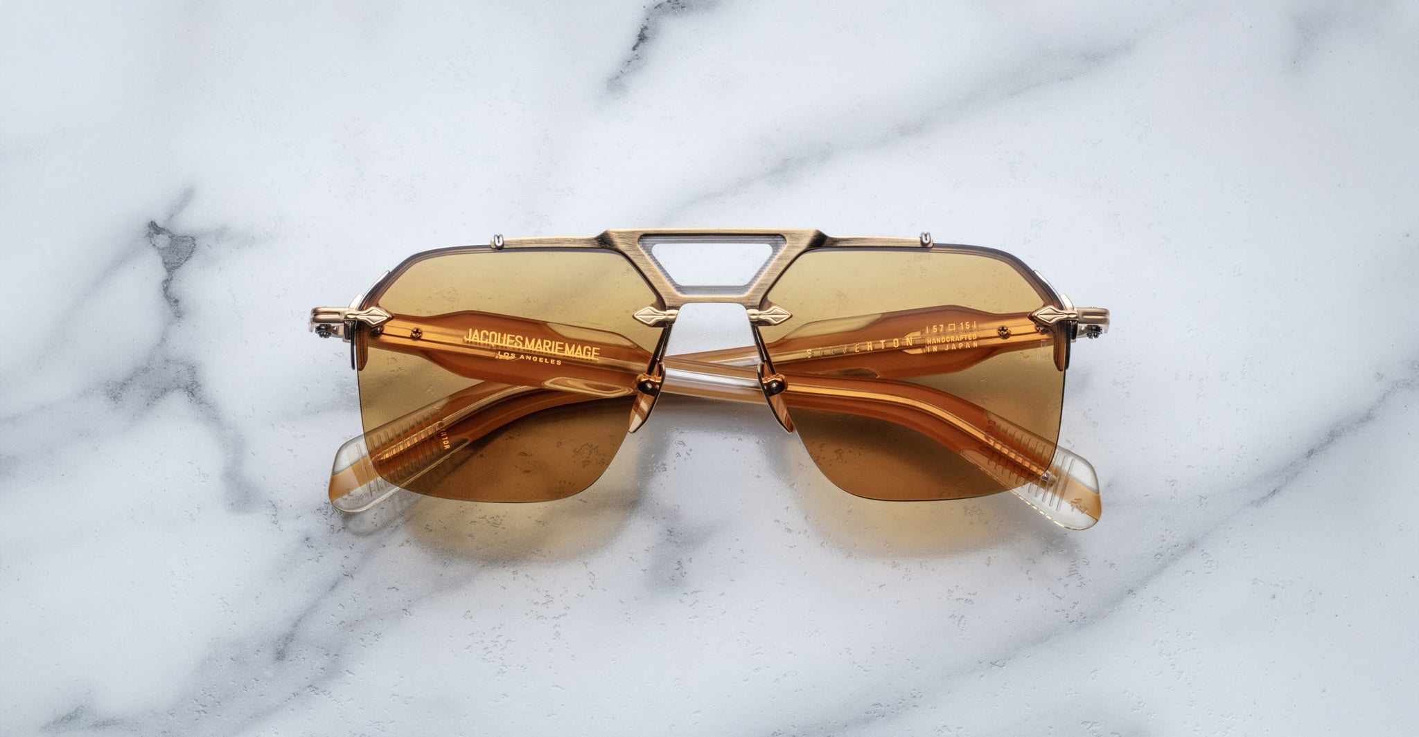 The Jacques Marie Mage Silverton sunglasses, featuring gold rims and brown tinted geometric lenses with visible text on the arms, are displayed on a white marble surface.