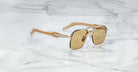The Jacques Marie Mage Silverton sunglasses, featuring a gold and black frame with metallic geometric accents on the arms and bridge and rectangular brown lenses, rest on a white marble surface.