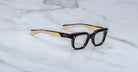 A pair of Jacques Marie Mage Stewart rectangular tortoiseshell eyeglasses with light inner arms rests on a white marble surface with soft gray veining.